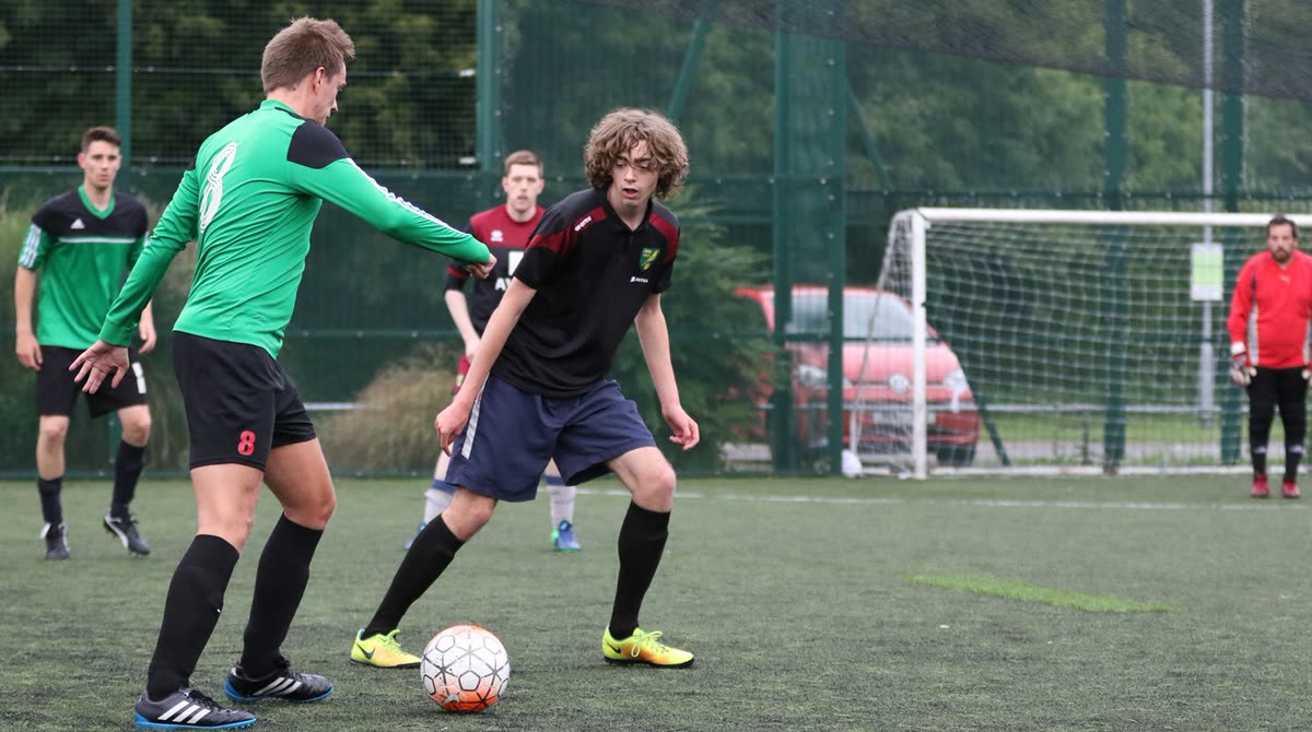 A game in play on a 3G pitch: a player in green shields the ball from an opponent while team-mates spread out behind them and a keeper watches from his goal.