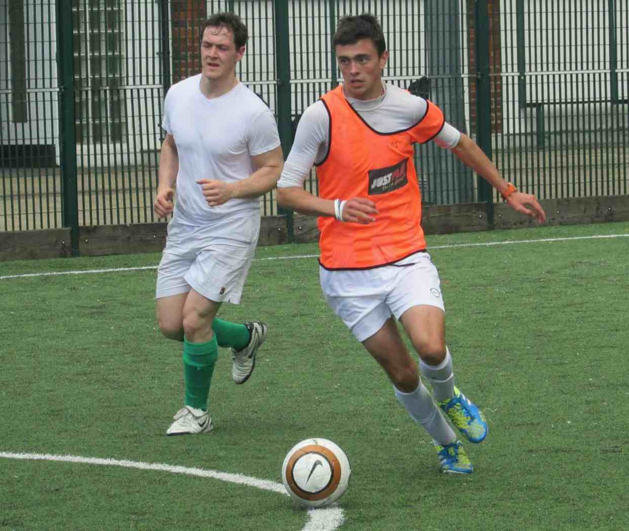 Two players running for the ball on a 3G pitch, one in an orange bib just ahead of the other.
